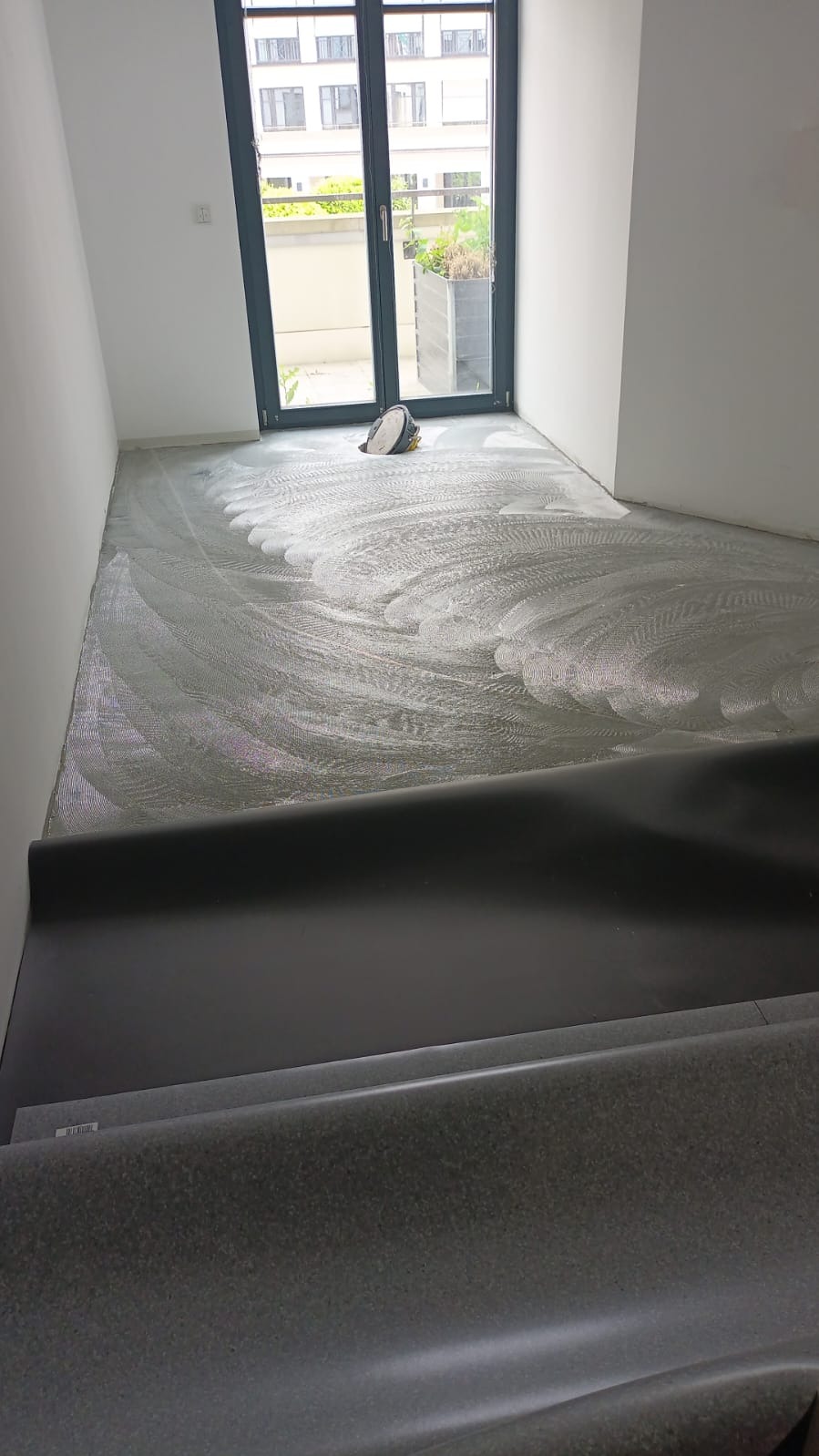 Przygotowanie podłoża pod nową szarą wykładzinę podłogową, widoczne resztki kleju i narzędzia w pustym pomieszczeniu z dużym oknem balkonowym.