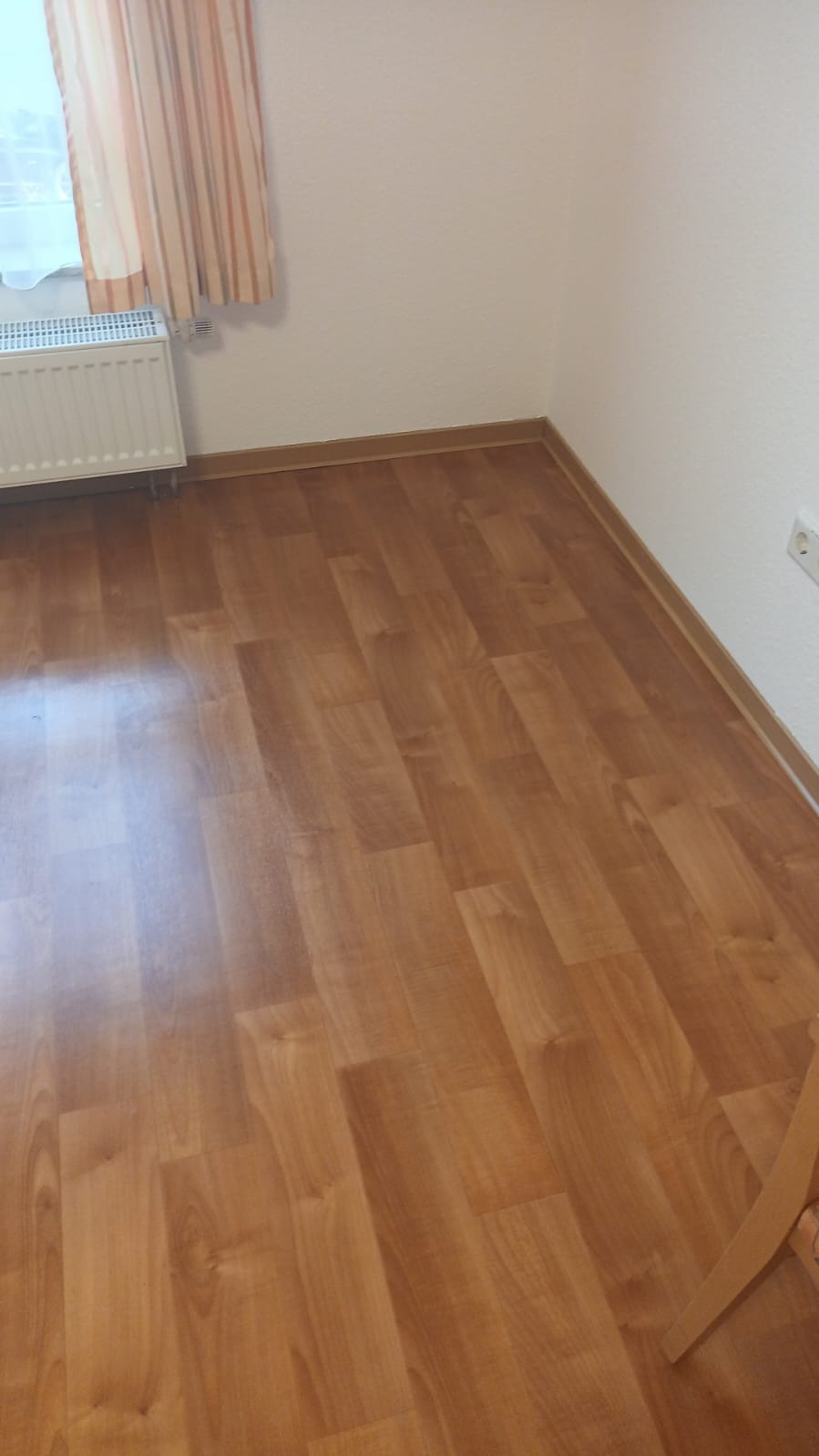 Jasnobrązowe panele podłogowe ułożone w pokoju, widoczna listwa przypodłogowa, grzejnik pod oknem i fragment zasłony w tle.
