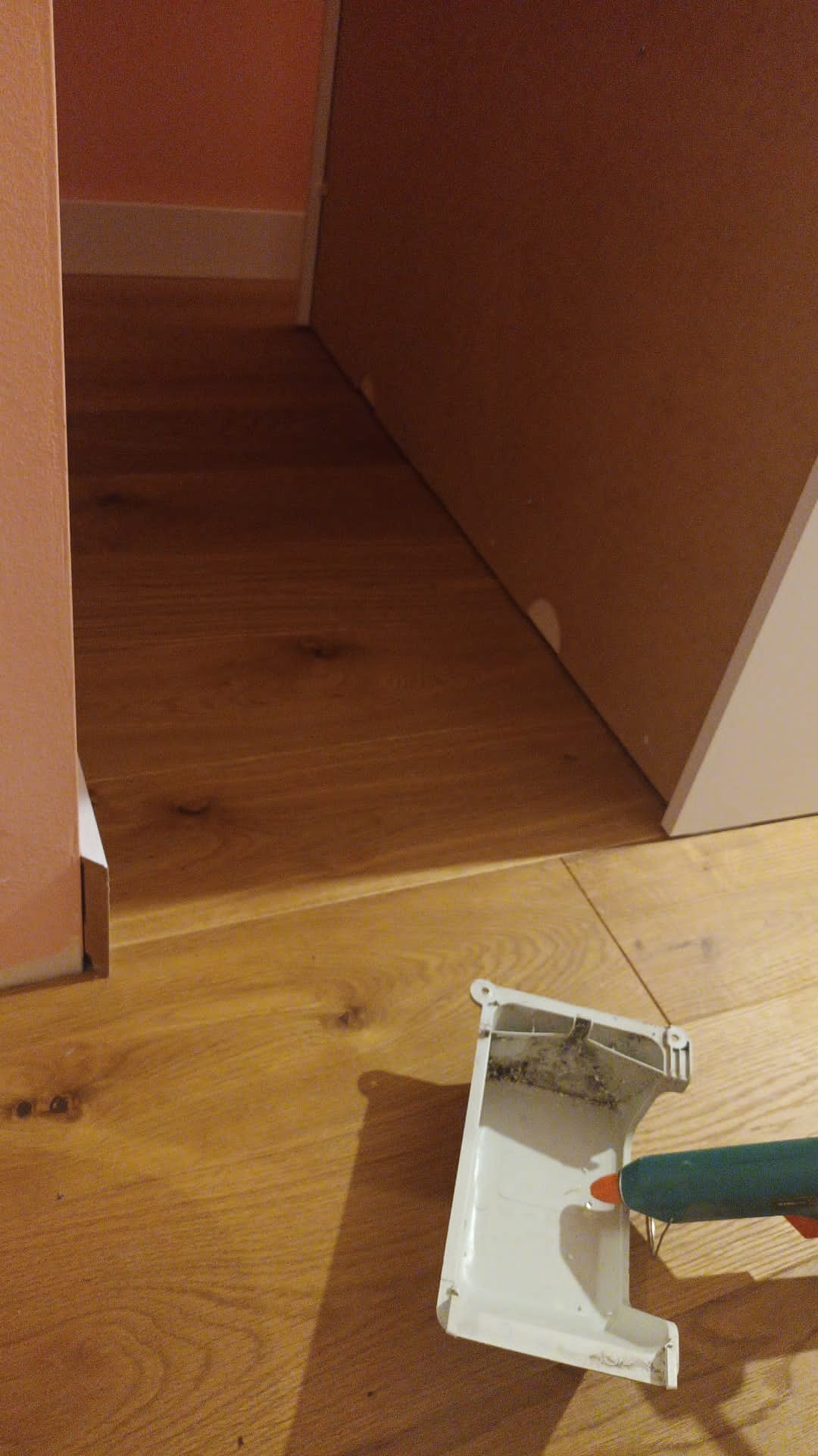 Fragment układania paneli pod ścianą, widoczna listwa przypodłogowa i pistolet do kleju z resztkami kleju na listwie.