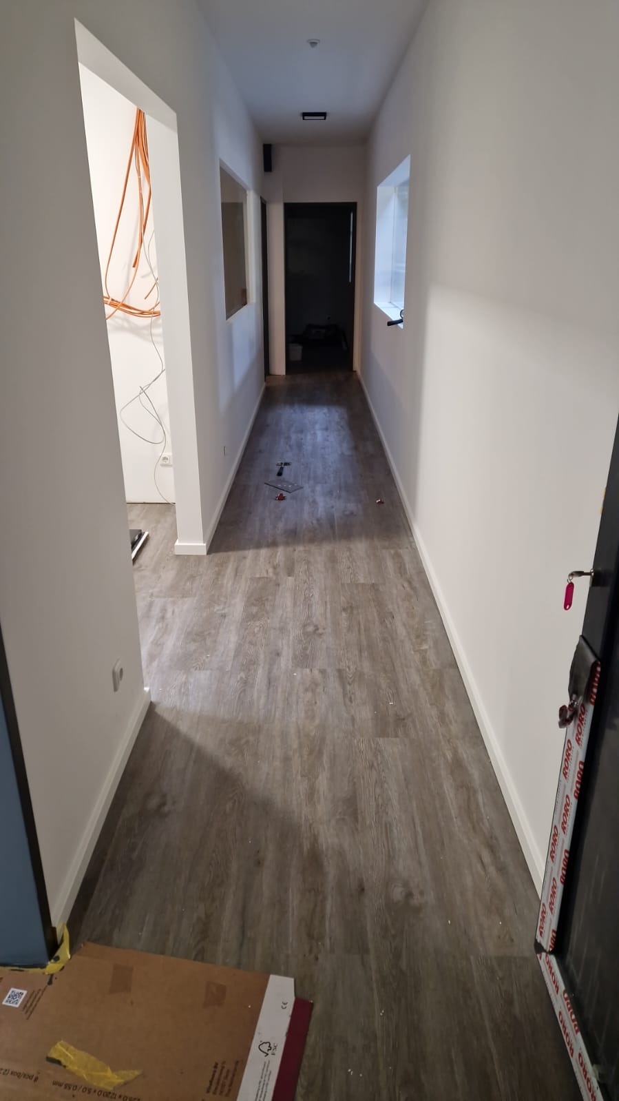 Korytarz po remoncie z nową podłogą z paneli imitujących drewno, widoczne narzędzia i materiały budowlane, białe ściany i przygotowane otwory na okna i instalacje elektryczne.