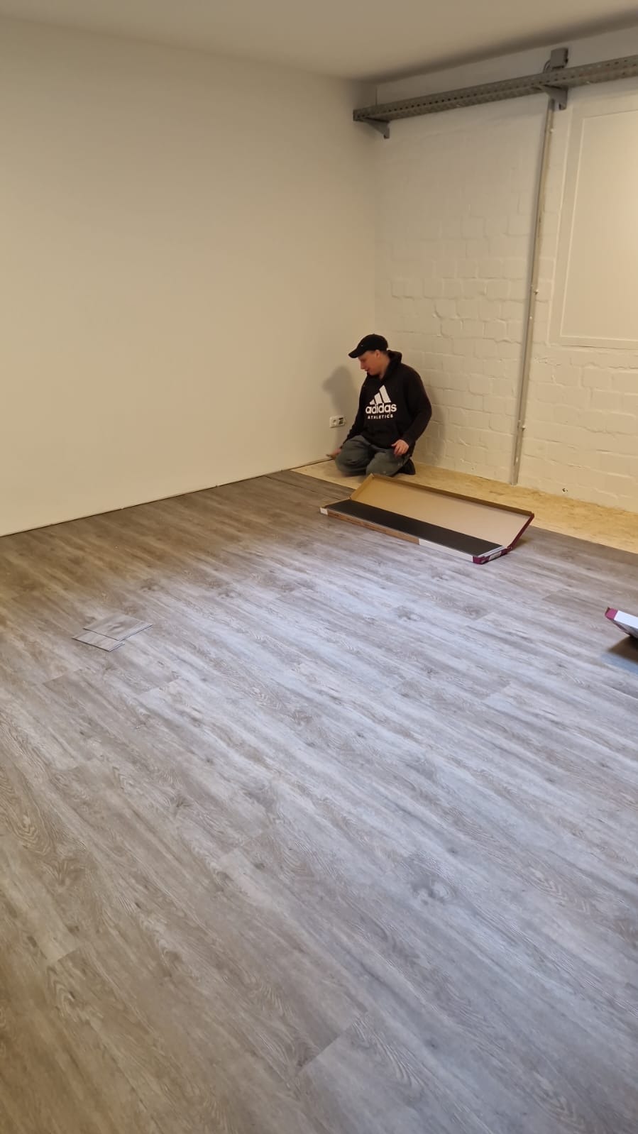 Pracownik w czarnej bluzie i czapce układa panele podłogowe w jasnym pomieszczeniu. Część podłogi jest już pokryta panelami o wzorze drewna, a obok leży otwarte opakowanie z panelami.