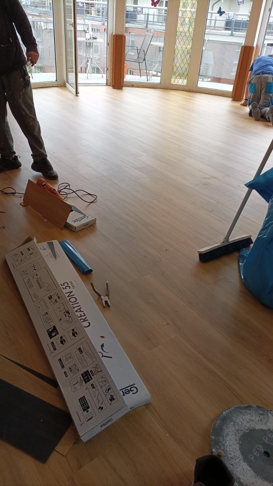 Montaż paneli podłogowych w jasnym pokoju z oknami balkonowymi, widoczne narzędzia, paczki paneli Gerflor Creation 55 i pracownicy podczas sprzątania po montażu.