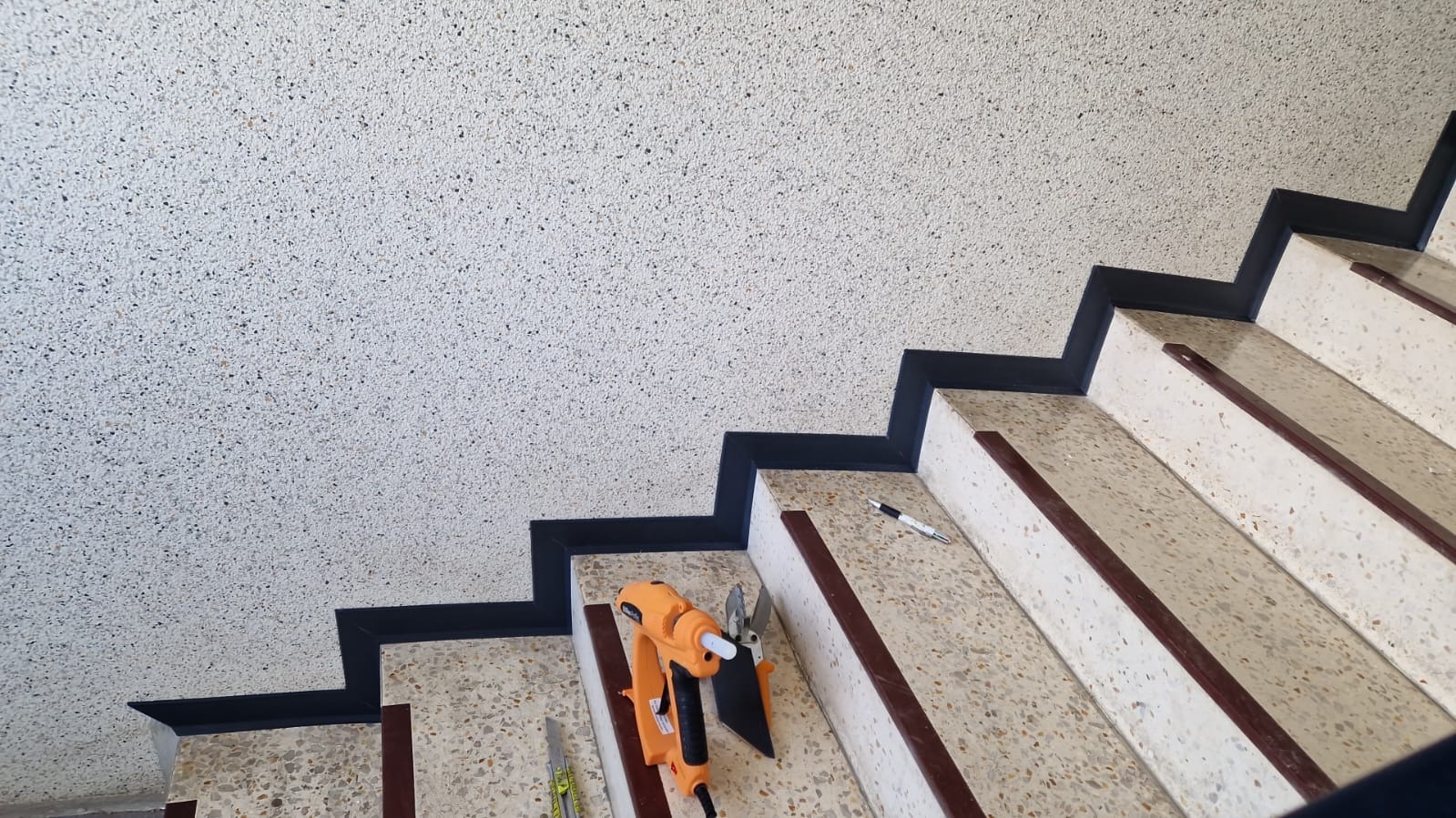 Schody z terazzo z czarnymi listwami przypodłogowymi, narzędzia do wykończenia, pistolet do kleju na gorąco i nóż do cięcia listew przypodłogowych na stopniu schodów.