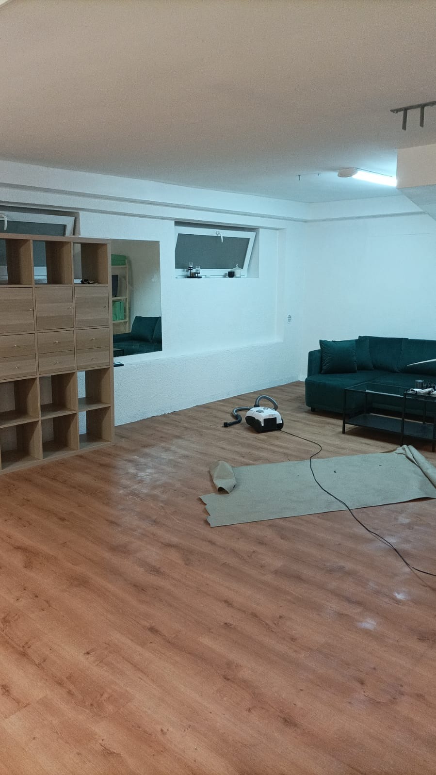 Salon po remoncie z nową podłogą z paneli, częściowo zakrytą ochronną folią, odkurzacz i regał na tle zielonej sofy.