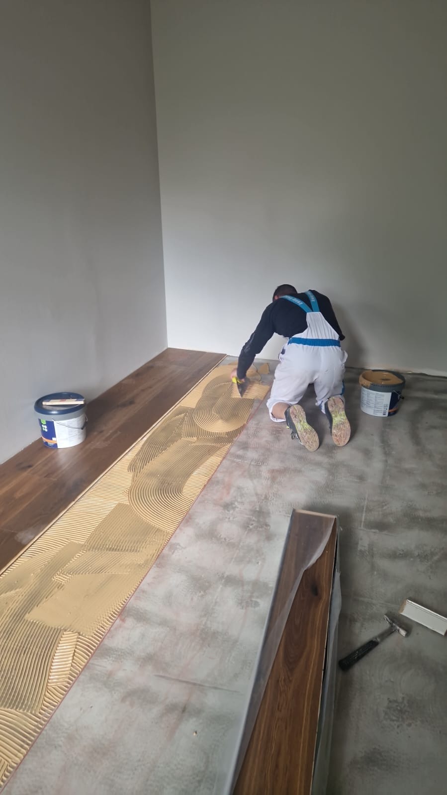 Parkieciarz rozprowadza klej szpachelką na podłożu pod panele podłogowe z widocznym wzorem w jodełkę, obok stoją puszki z klejem i leżą zapakowane panele.