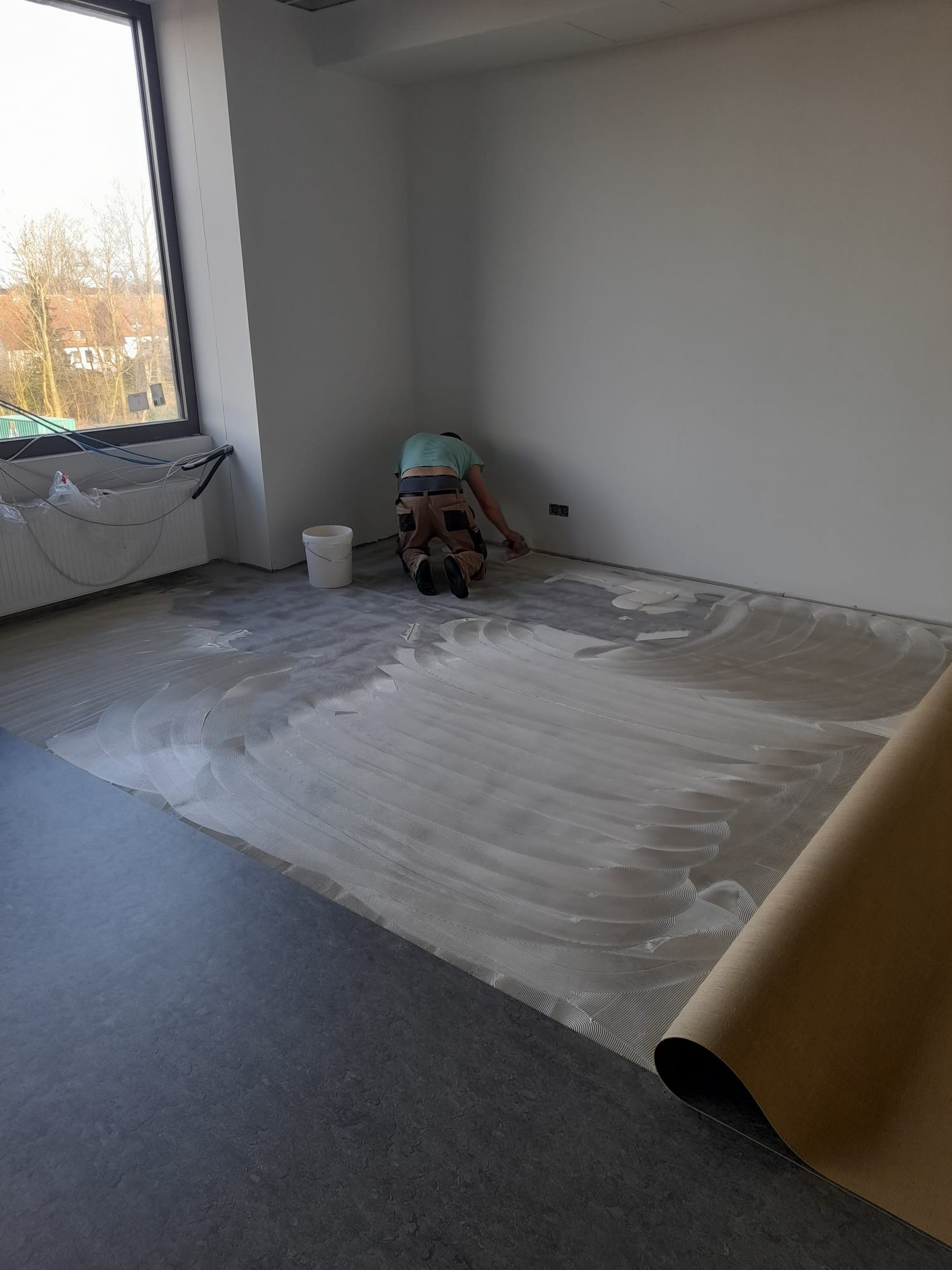 Pracownik na kolanach rozprowadza klej pod ciemną wykładzinę podłogową w jasnym pomieszczeniu, widoczny zwój wykładziny i wiadro z klejem w rogu pokoju.