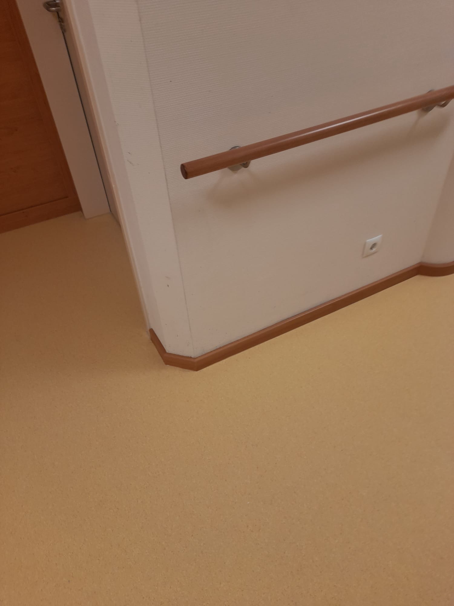 Korytarz z brązową poręczą przy ścianie, listwami przypodłogowymi w kolorze drewna i linoleum w odcieniach beżu.