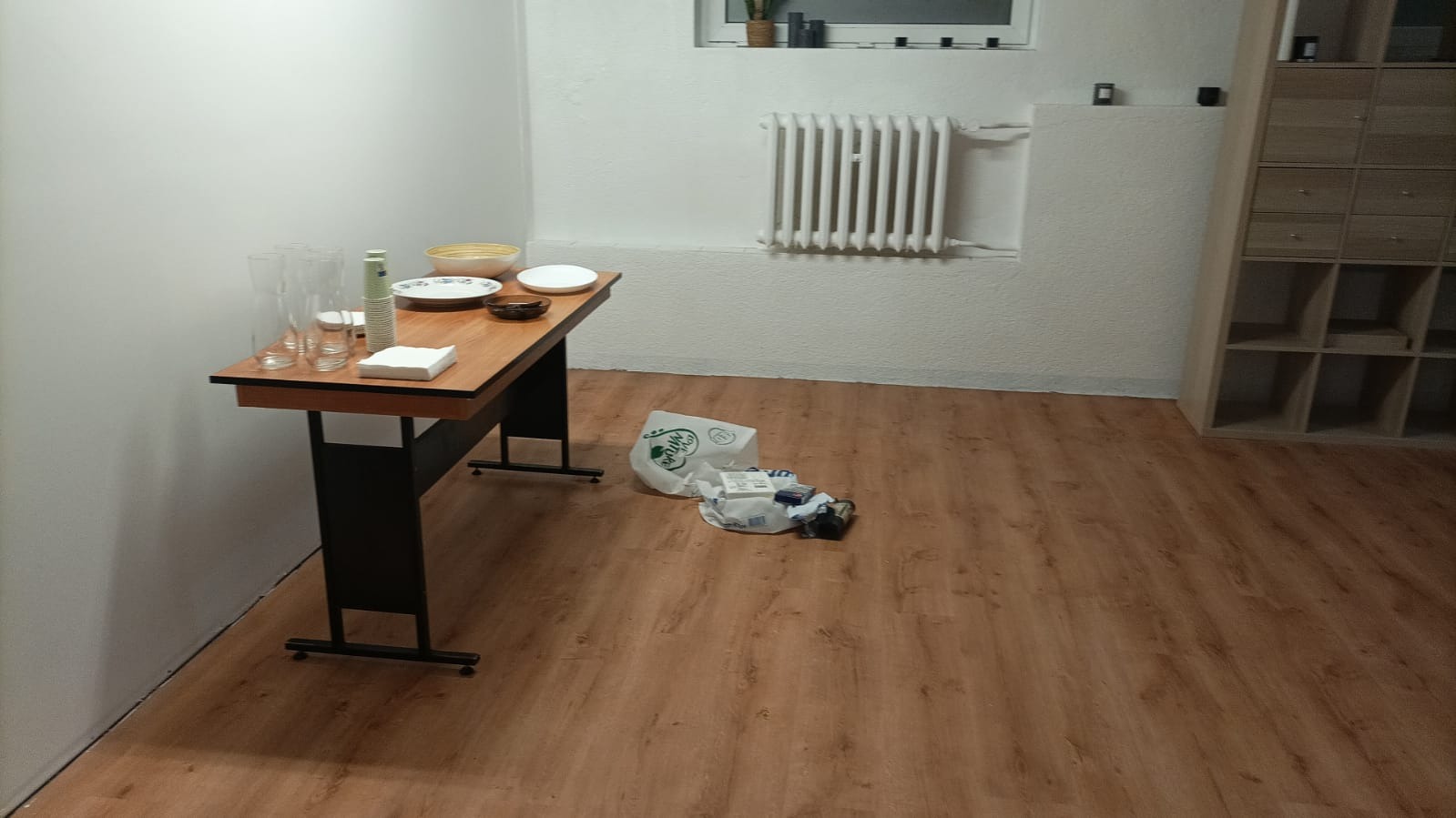 Wnętrze pokoju z panelami podłogowymi, stolikiem z naczyniami i torbami na podłodze. Widoczny grzejnik i regał z półkami.