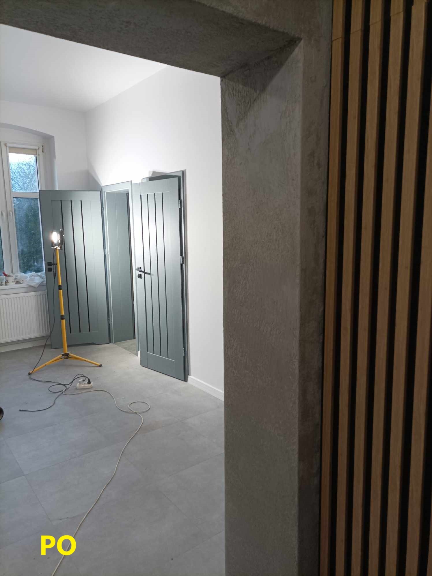 Wnętrze w trakcie remontu z dwiema szarymi, otwartymi parami drzwi, widoczna lampa robocza na trójnogu i fragment drewnianej ściany lamelowej, surowa ościeżnica.