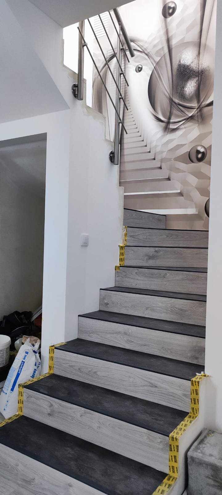 Schody wyłożone panelami imitującymi drewno w odcieniach szarości, zabezpieczone taśmą malarską, z abstrakcyjną tapetą na ścianie i metalową balustradą.