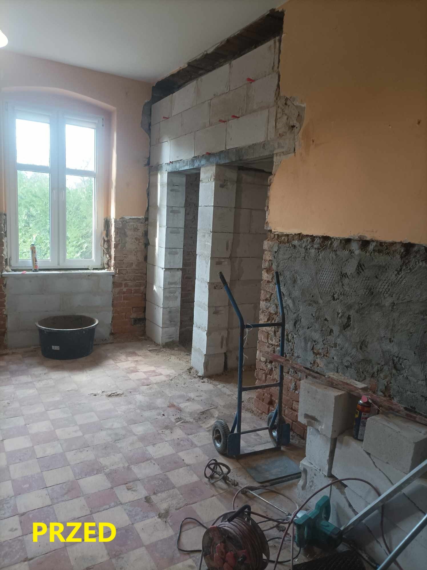 Wnętrze pokoju w trakcie zmiany aranżacji, widoczne niedokończone ściany z cegły i bloczków, w tle okno, na podłodze narzędzia i wózek transportowy, napis 'PRZED' na dole kadru.
