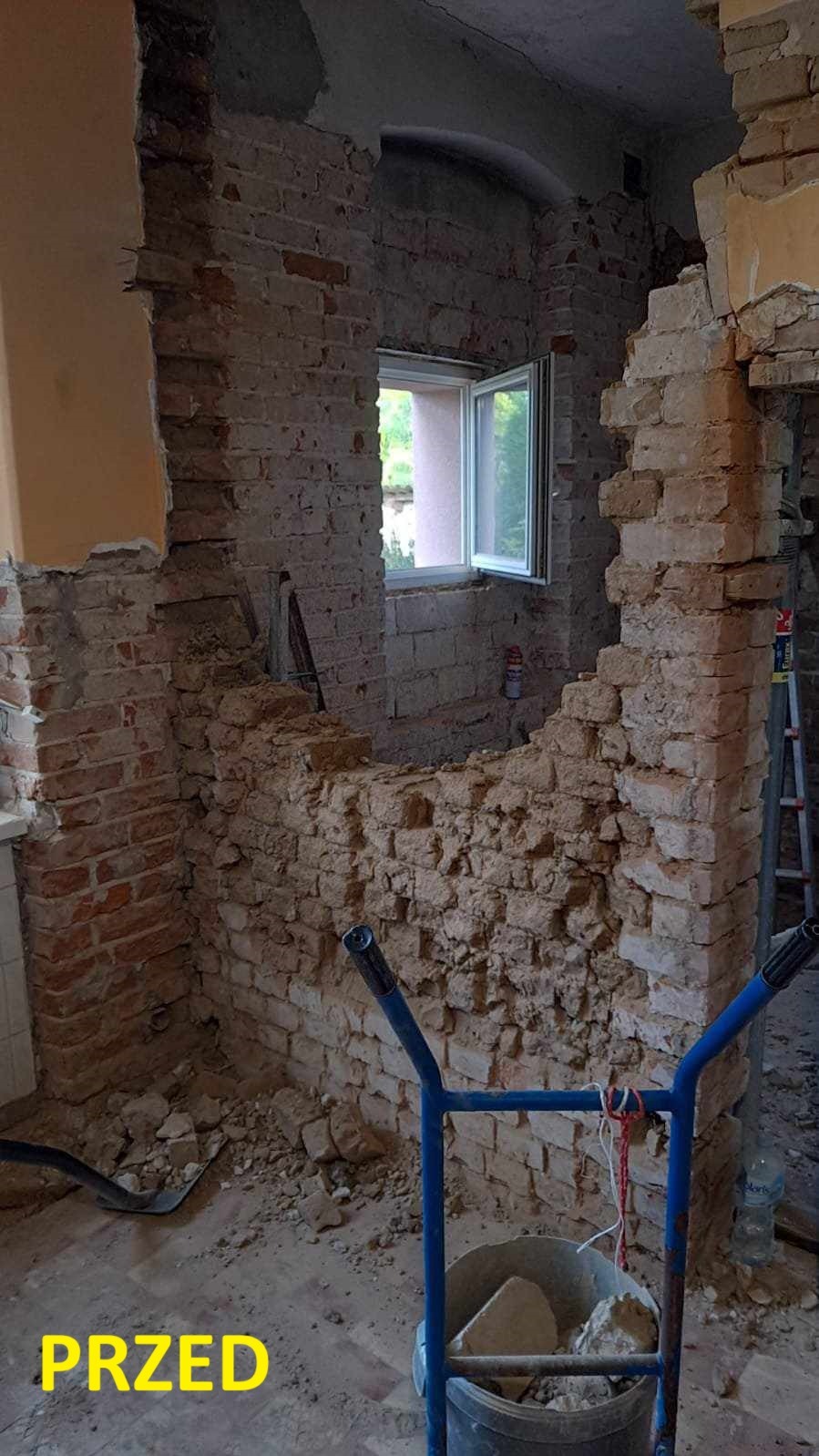 Wnętrze w trakcie demontażu ściany z cegły, widoczne odsłonięte cegły, gruz, okno z widokiem na zieleń i wózek budowlany z wiadrem pełnym odłamków.