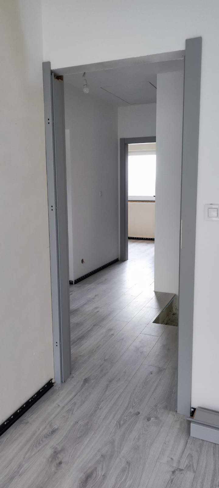 Szare panele podłogowe ułożone w jasnym wnętrzu, widoczne ościeżnice drzwi, surowy stan wykończenia