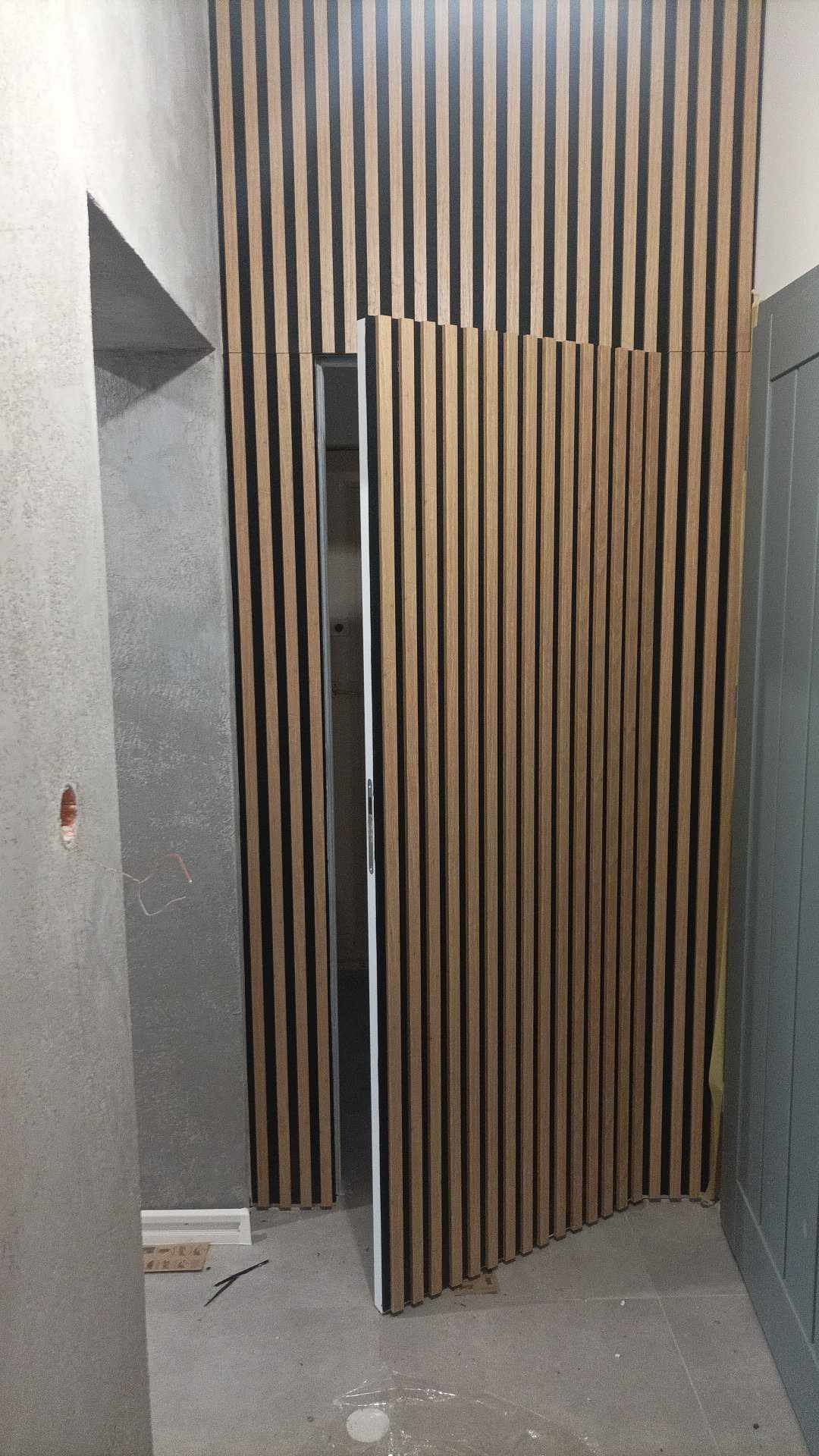 Montaż dekoracyjnych paneli ściennych z drewnianymi listwami na czarnym tle, częściowo zasłaniających otwarte drzwi w surowym wnętrzu.