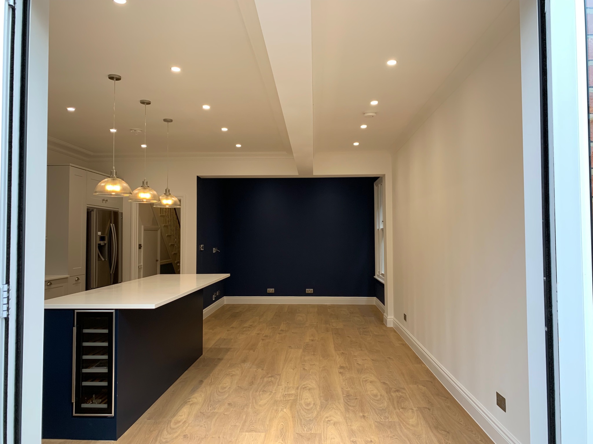 Otwarta przestrzeń po remoncie mieszkania: jasna kuchnia z wyspą, ciemnoniebieską ścianą akcentującą oraz podłogą z paneli imitujących drewno, widok z otwartych drzwi balkonowych.