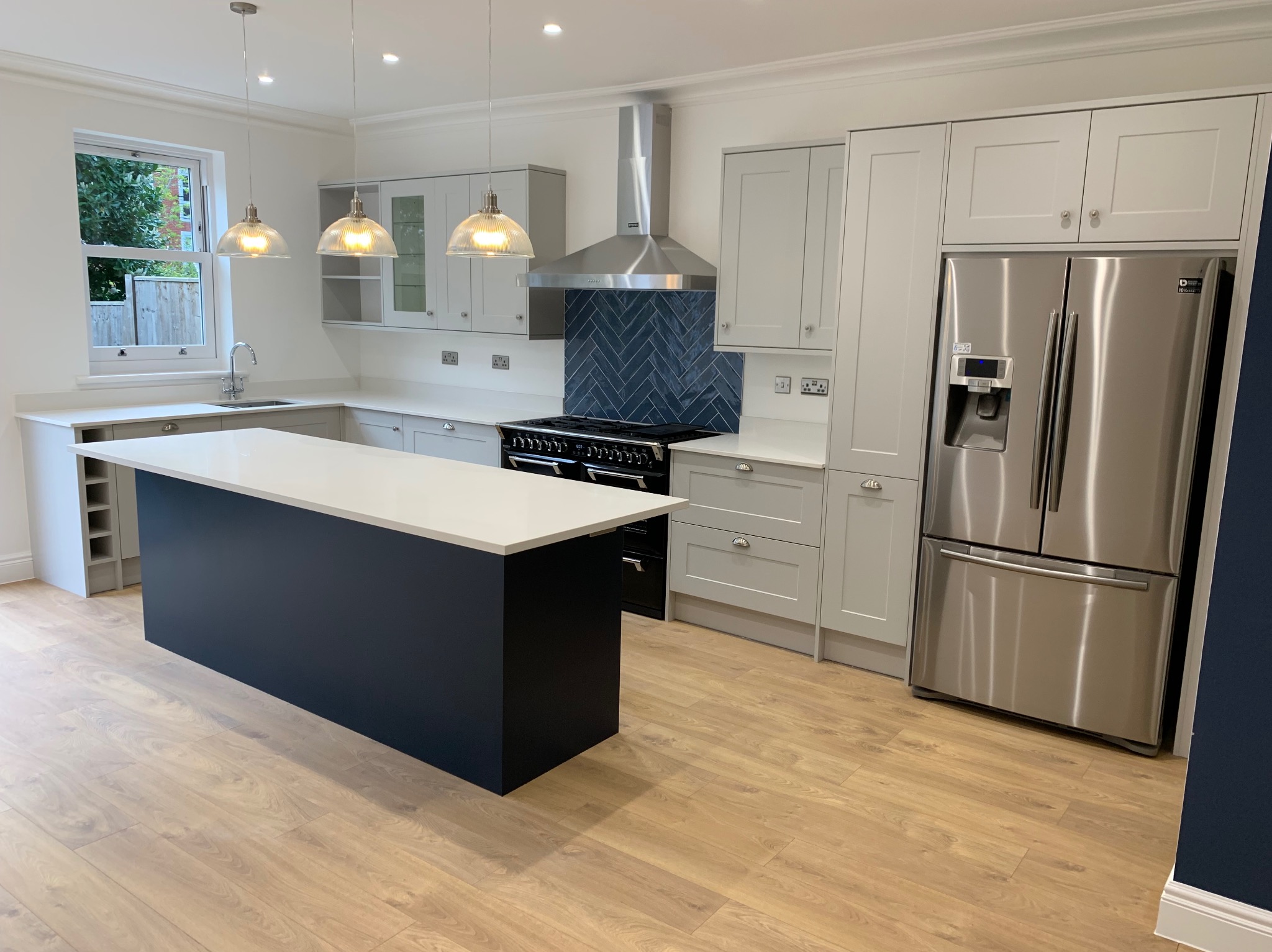 Jasna kuchnia z ciemnogranatową wyspą, drewnianą podłogą, szarymi szafkami i stalową lodówką. Nad wyspą wiszą trzy szklane lampy. Ściana nad kuchenką wykończona granatową cegłą w jodełkę.