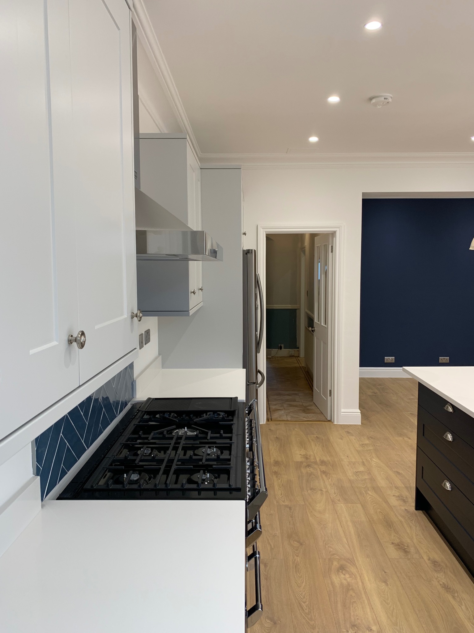 Nowoczesna kuchnia z białymi szafkami, niebieskimi płytkami w jodełkę nad kuchenką gazową i drewnianą podłogą, widok na odnowione pomieszczenie z granatową ścianą.