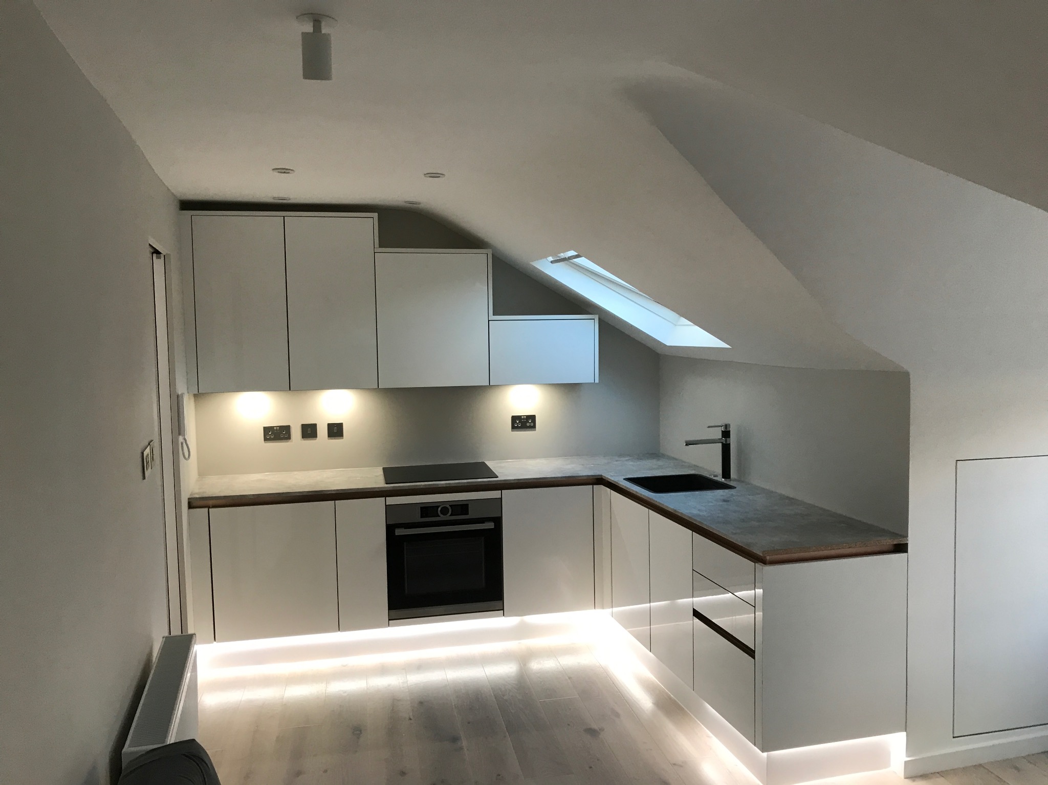 Nowoczesna, minimalistyczna kuchnia w odcieniach bieli z podświetleniem LED pod szafkami, umieszczona na poddaszu ze skośnym sufitem i oknem dachowym.