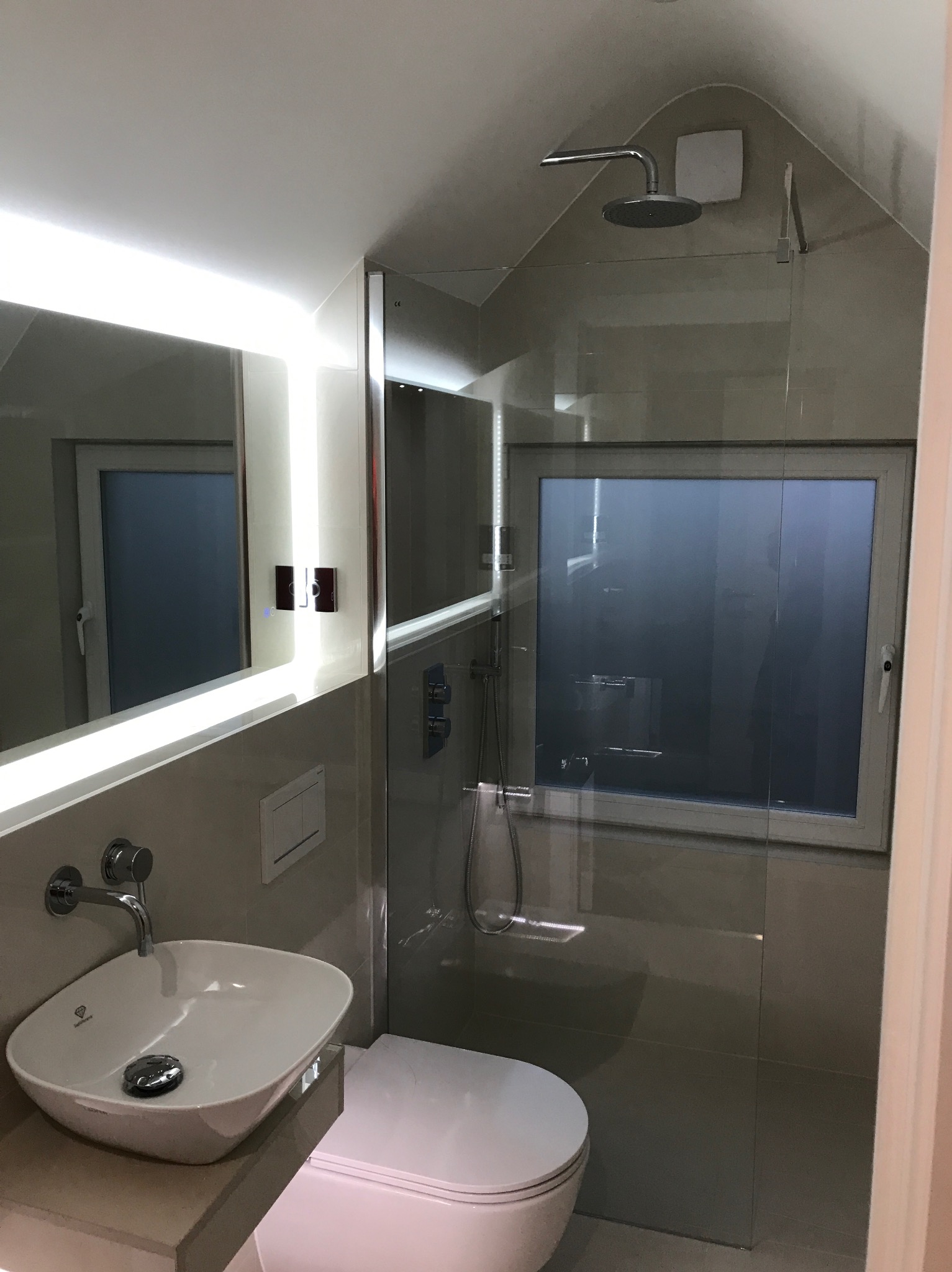 Nowoczesna łazienka z podświetlanym lustrem, kabiną prysznicową typu walk-in i wiszącą toaletą w odcieniach szarości, widoczna wentylacja pod sufitem.