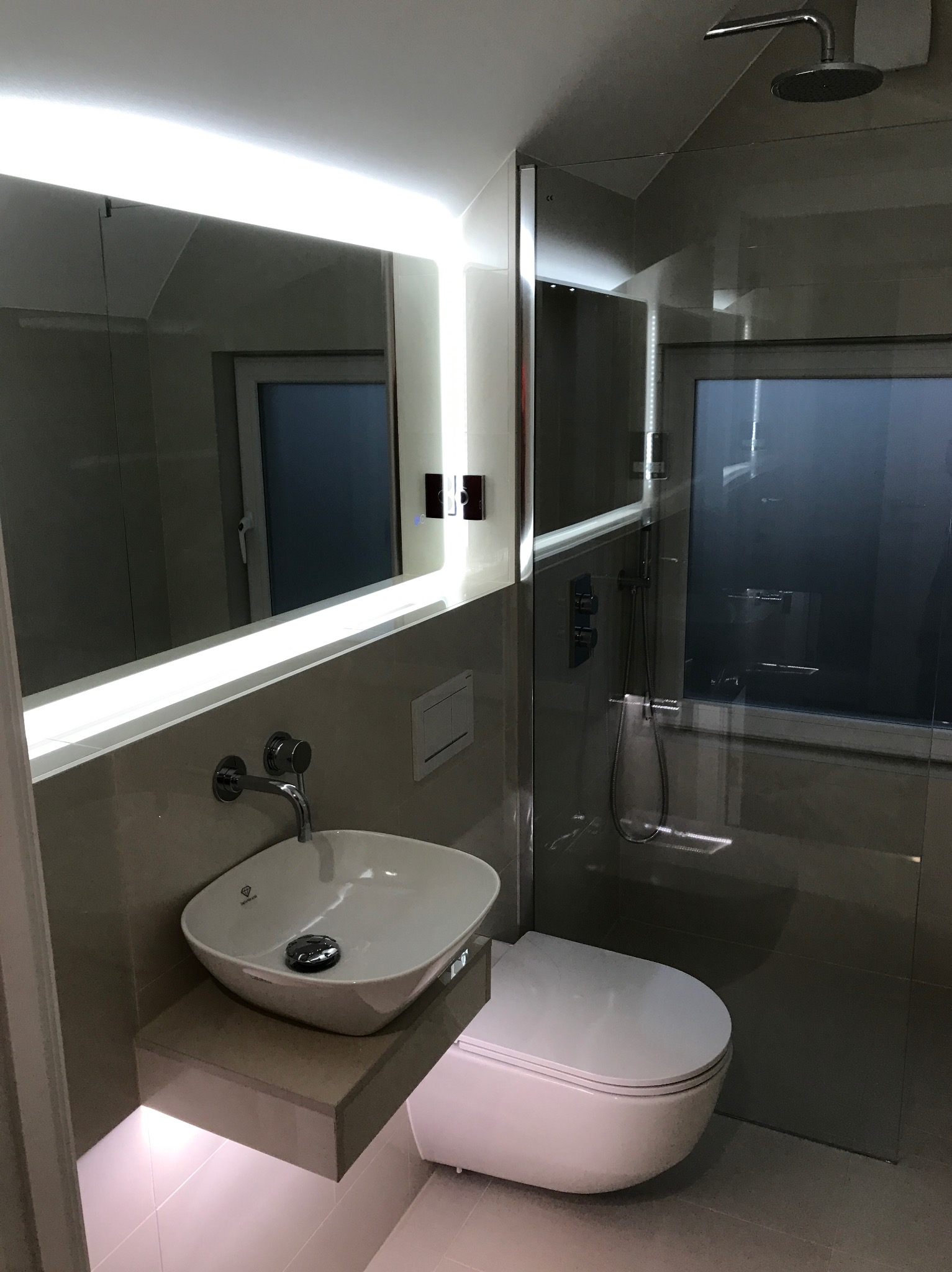 Nowoczesna łazienka z podświetlanym lustrem, wiszącą umywalką na kamiennym blacie z subtelnym podświetleniem LED oraz kabiną prysznicową ze szklaną ścianą i deszczownicą.