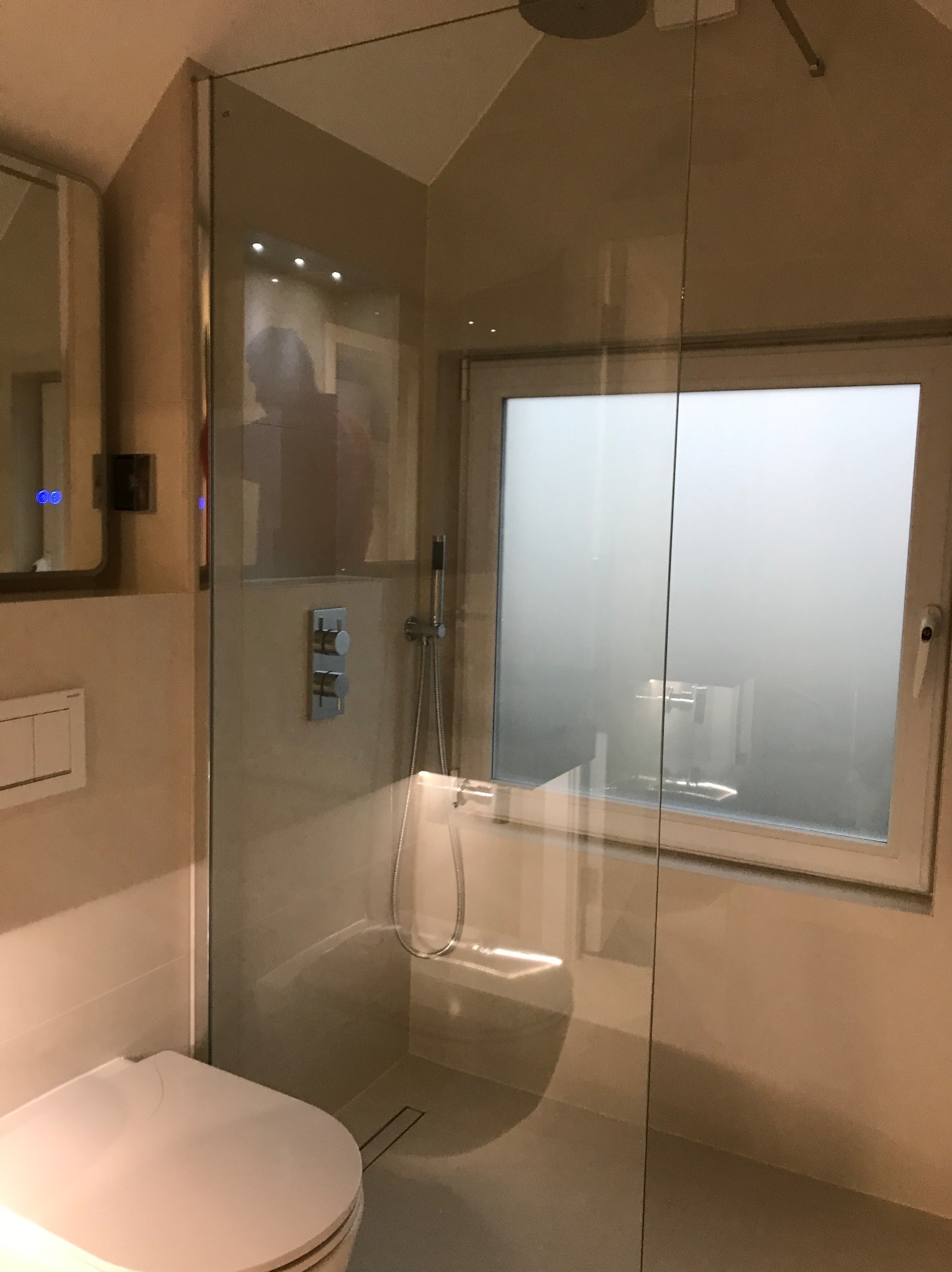 Nowoczesna łazienka z kabiną prysznicową typu walk-in, widoczne okno z mleczną szybą, minimalistyczna armatura i toaleta w jasnych odcieniach beżu.