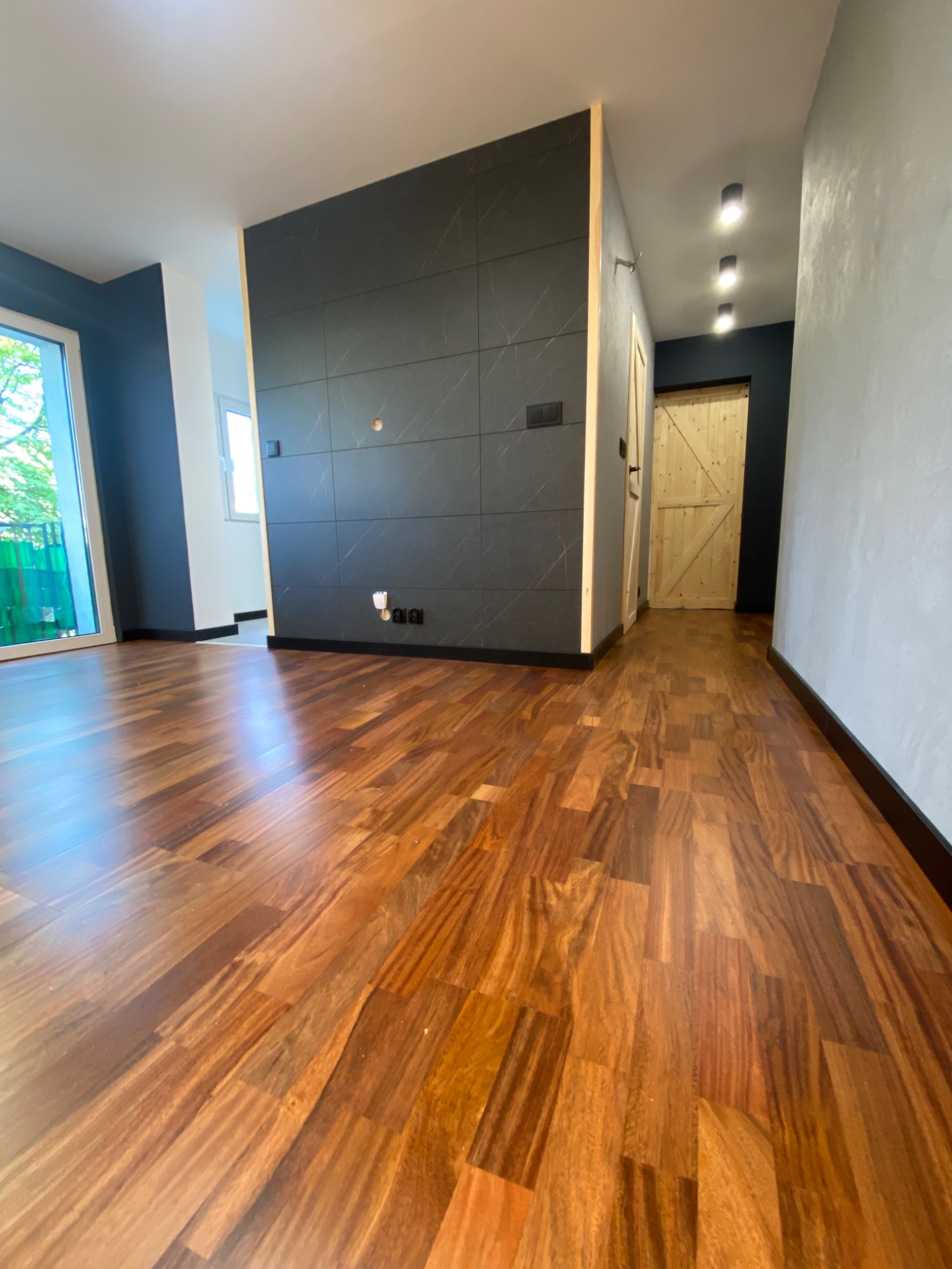 Wnętrze mieszkania po remoncie: parkiet z egzotycznego drewna, czarna ściana z płyt imitujących marmur, widoczne gniazdka elektryczne i otwór na kontakt, drewniane drzwi w stylu rustykalnym.