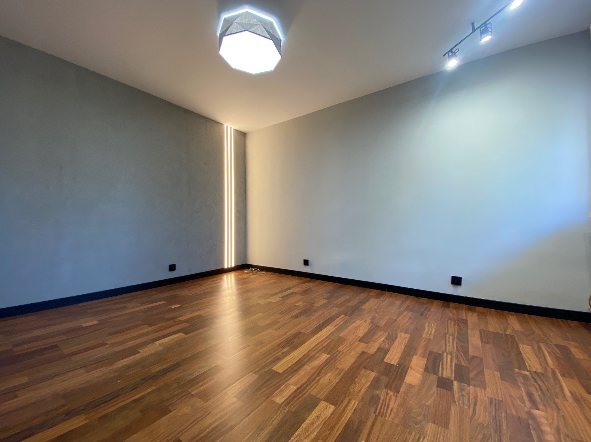 Pusty pokój z parkietem w jodełkę, szarą ścianą z pionowym oświetleniem LED w rogu, nowoczesną lampą sufitową i reflektorami