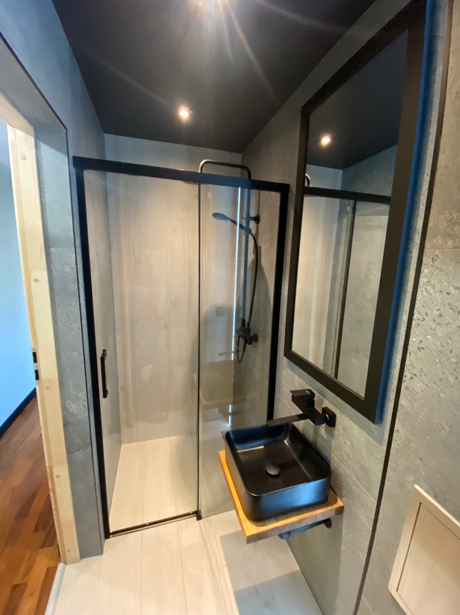 Nowoczesna łazienka z kabiną prysznicową typu walk-in z czarnymi profilami, czarną armaturą i umywalką nablatową na drewnianym blacie, szare płytki na ścianach i suficie.