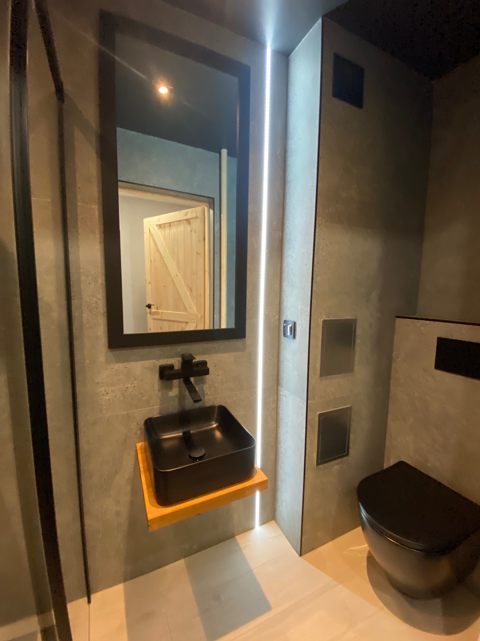 Nowoczesna łazienka z czarną umywalką nablatową na drewnianym blacie, czarną armaturą i wiszącą toaletą w kolorze czarnym, odbicie drewnianych drzwi w lustrze.