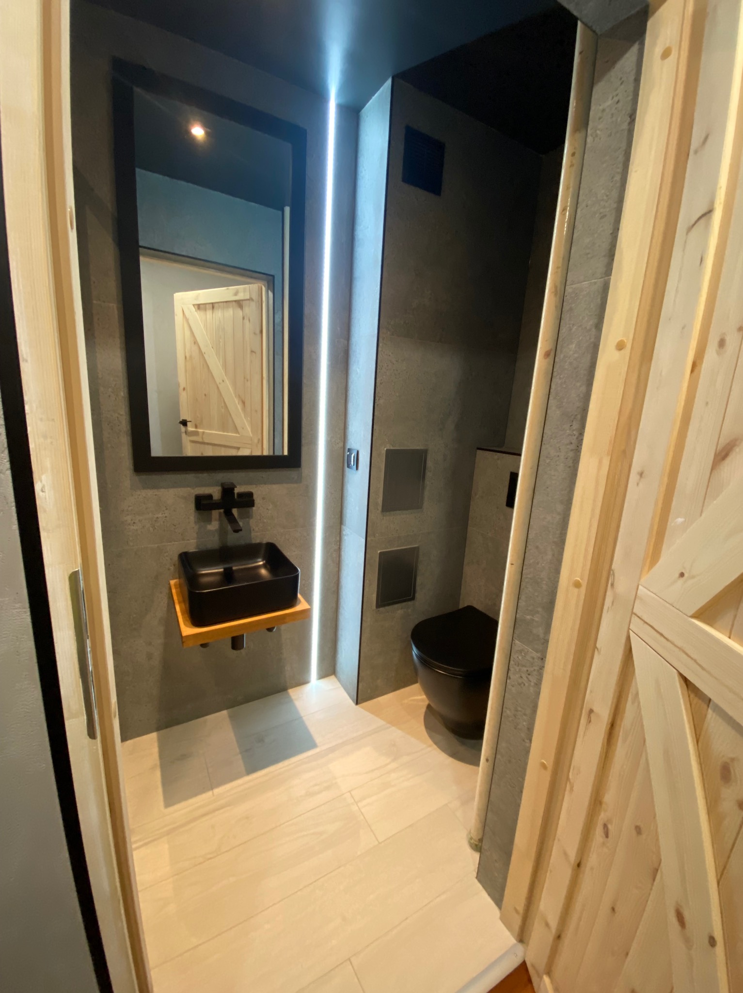 Nowoczesna łazienka z czarną armaturą, umywalką w kształcie kostki i toaletą, oświetleniem LED oraz drewnianymi drzwiami w stylu rustykalnym.