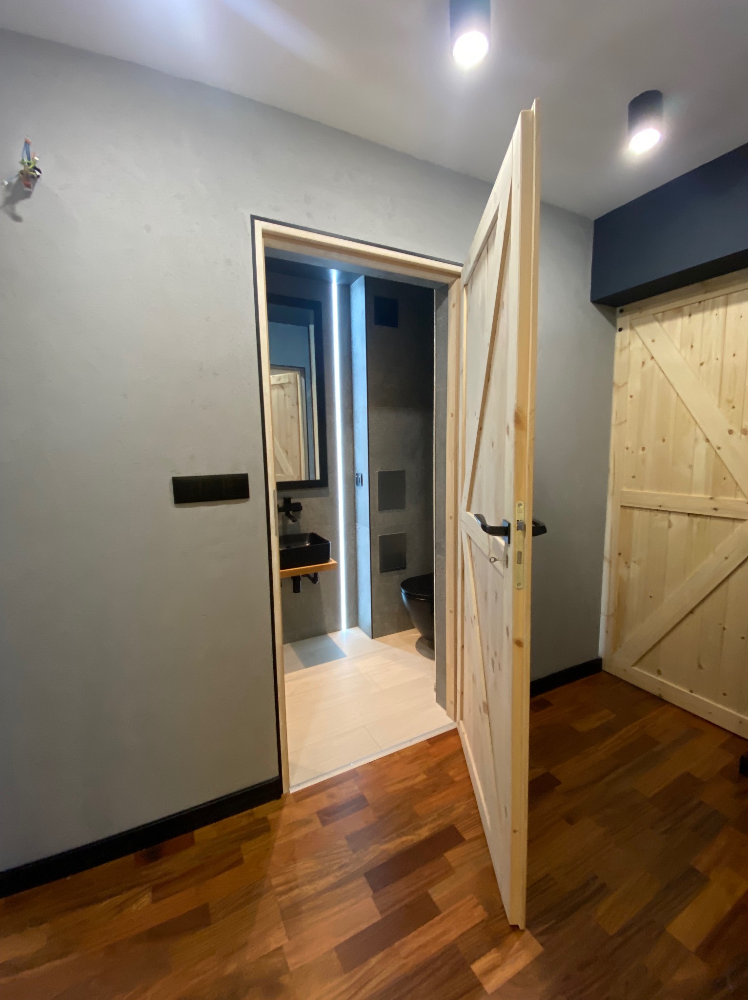 Otwarta drewniana drzwi do szarej łazienki z czarną umywalką i toaletą, widok na parkiet z desek ułożonych w jodełkę.