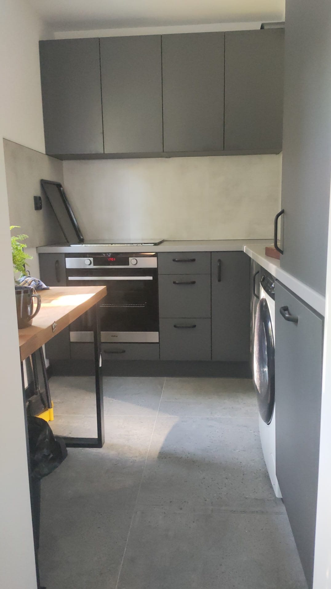 Nowoczesna kuchnia w odcieniach szarości z drewnianym blatem stołu, widoczna pralka i piekarnik.