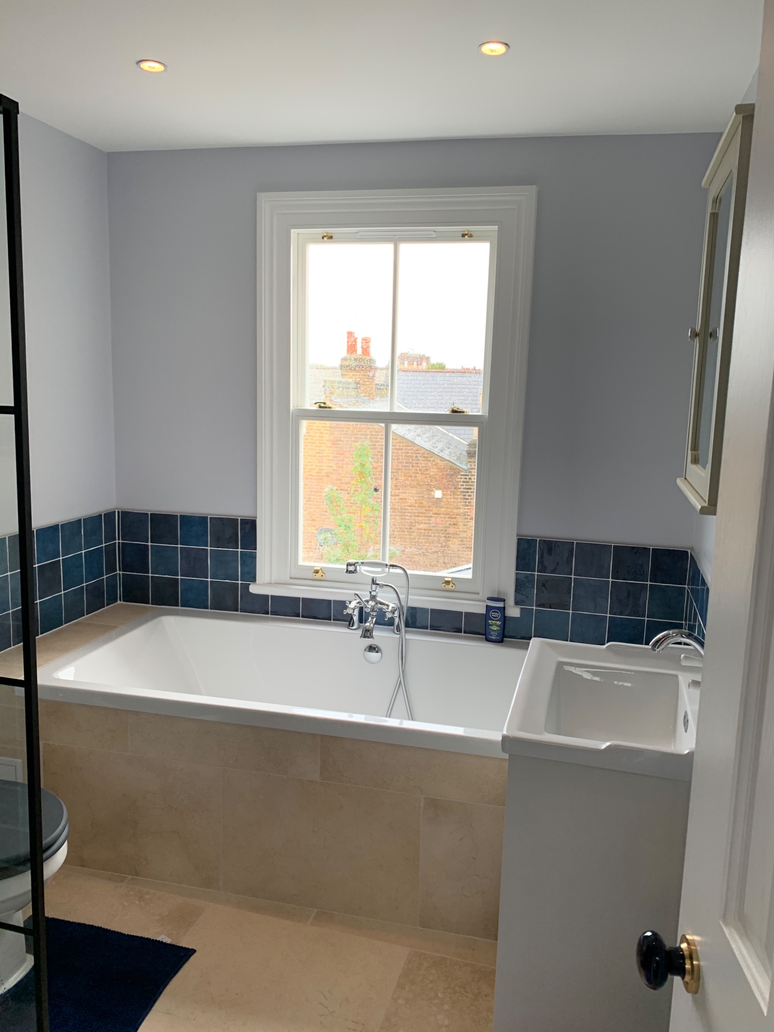 Jasna łazienka z wanną pod oknem, wykończona beżowymi płytkami i ciemnoniebieskimi kaflami ściennymi, z widoczną zabudową umywalki i fragmentem kabiny prysznicowej.