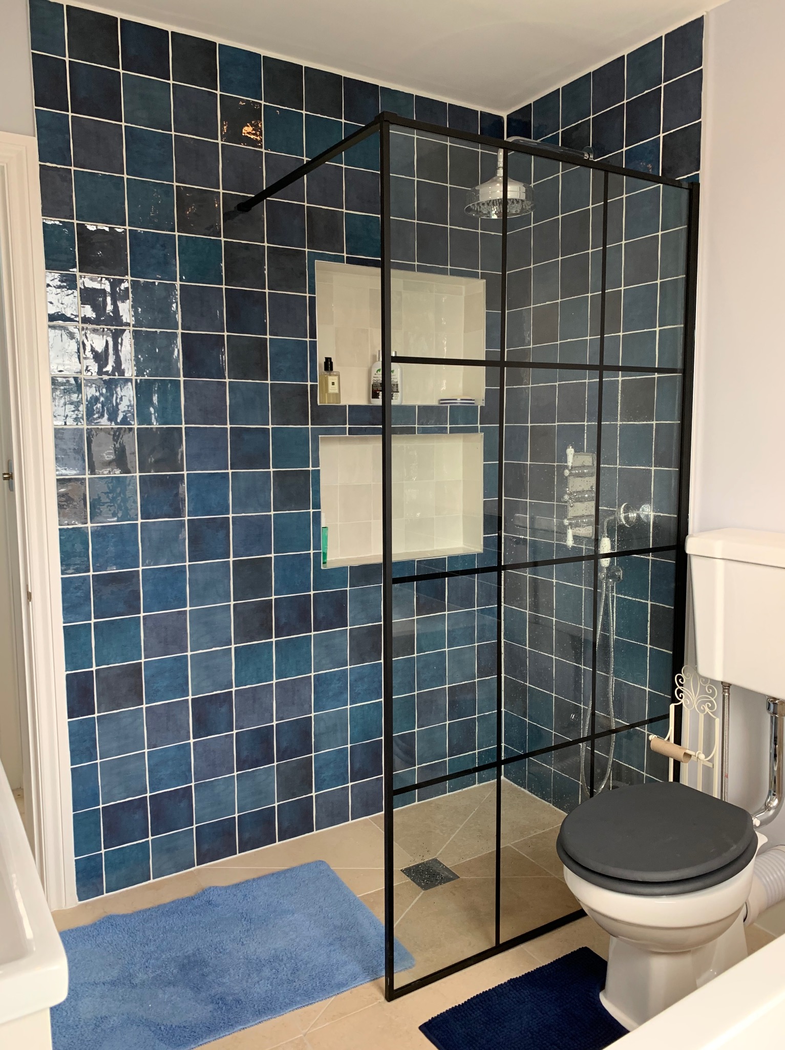 Wyremontowana łazienka z kabiną prysznicową typu walk-in z czarnymi profilami, niebieskimi kwadratowymi płytkami na ścianach, wbudowanymi półkami i beżową podłogą.