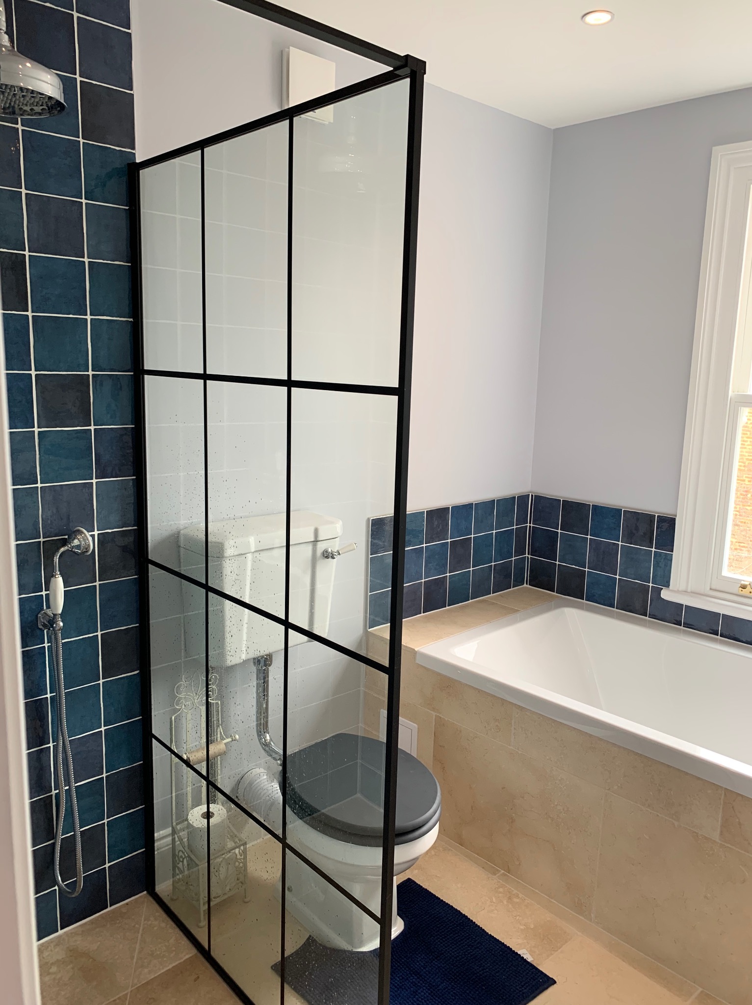 Nowoczesna łazienka z kabiną prysznicową typu walk-in z czarnymi szprosami, niebieskimi płytkami w strefie prysznica i nad wanną, beżową podłogą i białymi sanitariatami.