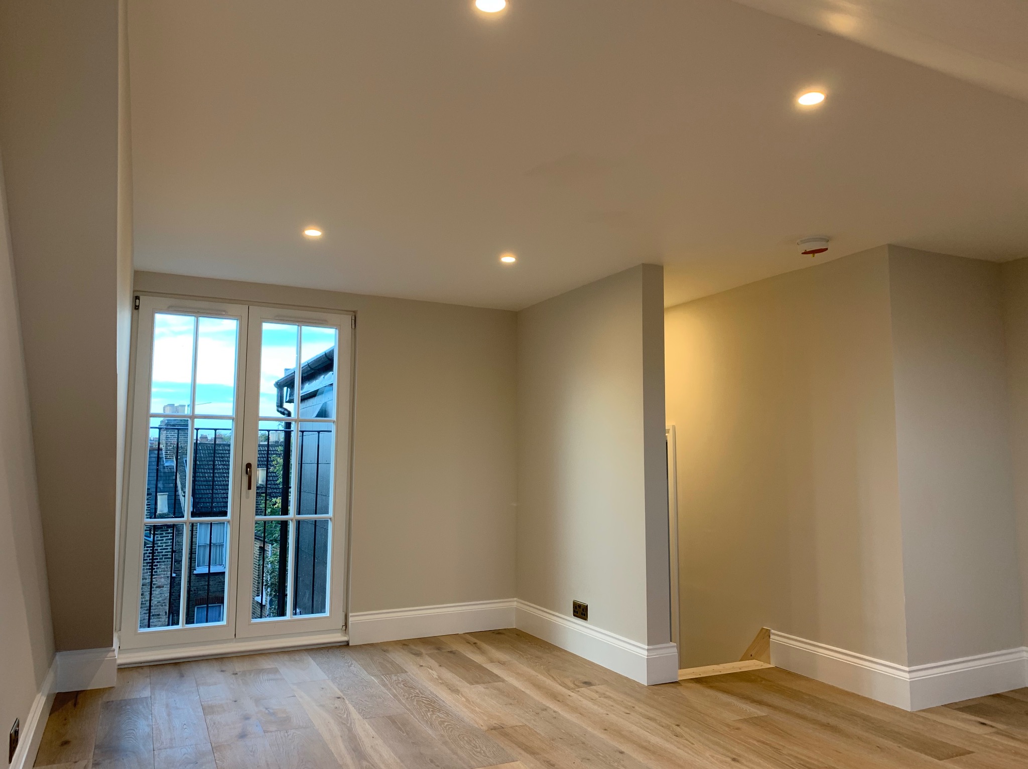 Jasne, przestronne wnętrze poddasza z drewnianą podłogą, oknem balkonowym z widokiem na zabudowę miejską i oświetleniem punktowym w suficie.