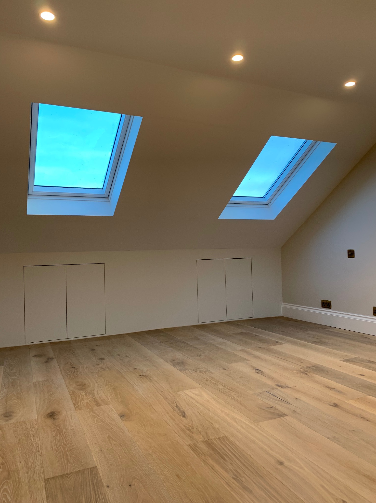 Jasne, przestronne poddasze z drewnianą podłogą, dwoma oknami dachowymi z widokiem na niebo, wbudowanymi szafkami i punktowym oświetleniem.