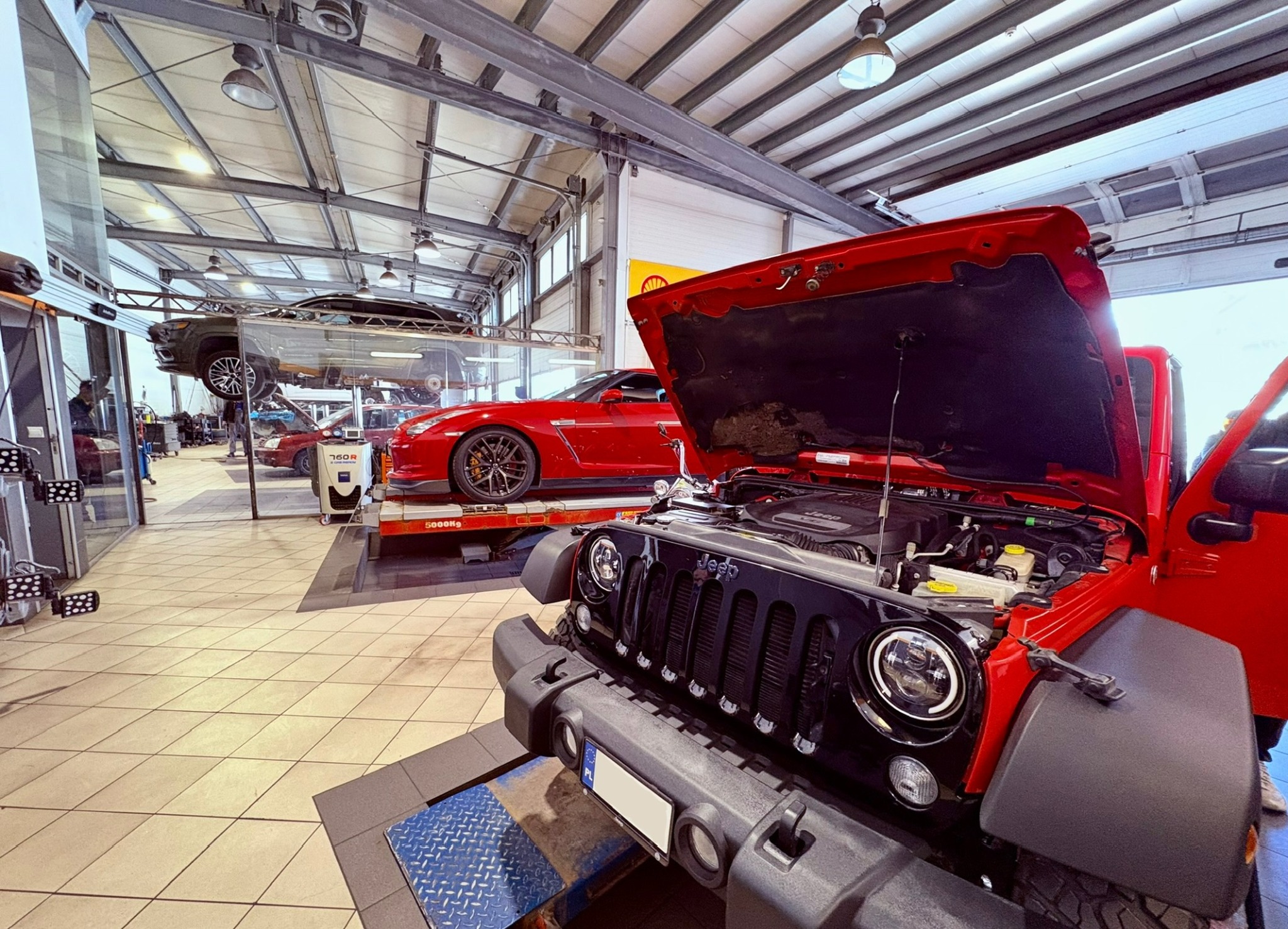 Czerwony Jeep Wrangler z otwartą maską w warsztacie samochodowym, obok czerwony Nissan GT-R na podnośniku. W tle inne auta na podnośnikach i sprzęt diagnostyczny.