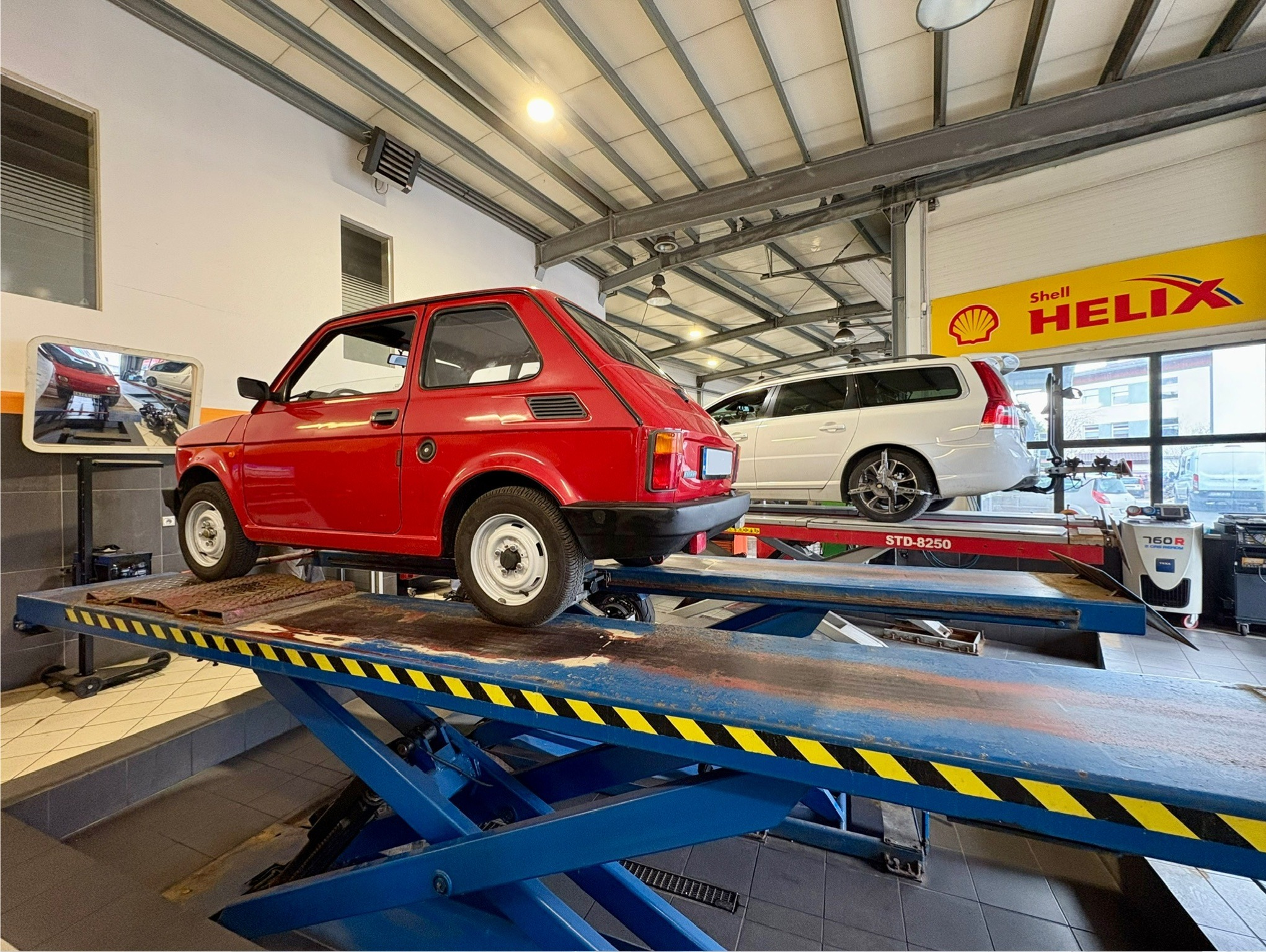 Czerwony Fiat 126p na podnośniku w warsztacie samochodowym. W tle biały samochód na drugim podnośniku i logo Shell Helix. Widok z boku, oświetlenie warsztatowe.