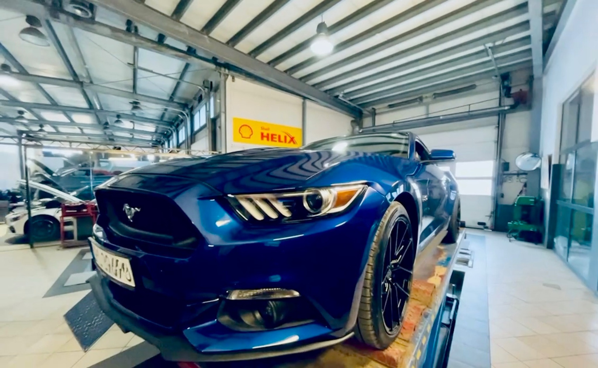 Niebieski Ford Mustang na podnośniku w warsztacie. Widoczne logo Shell Helix, oświetlenie sufitowe i inne auta w tle. Diagnostyka zawieszenia i naprawa.