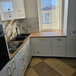 Grzegorz Wierzbicki - Nowoczesna kuchnia z białymi szafkami i złotymi uchwytami, marmurowym backsplashiem i drewnianym blatem, widziana pod kątem z brązową podłogą w romby.