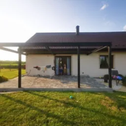 Nowoczesna, czarna konstrukcja zadaszenia tarasu przylegająca do domu z brązowym dachem, widok na trawnik i suszarkę na pranie.