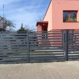 Nowoczesna, pozioma brama wjazdowa w kolorze grafitowym z furtką, na tle różowego domu jednorodzinnego, ułożona kostka brukowa.