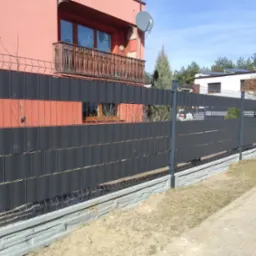 Nowoczesne ogrodzenie panelowe z czarnymi wypełnieniami na betonowej podmurówce, na tle różowego domu z balkonem i zielenią