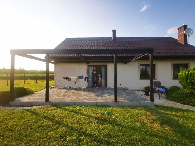 Nowoczesna, czarna konstrukcja zadaszenia tarasu przylegająca do domu z brązowym dachem, widok na trawnik i suszarkę na pranie.
