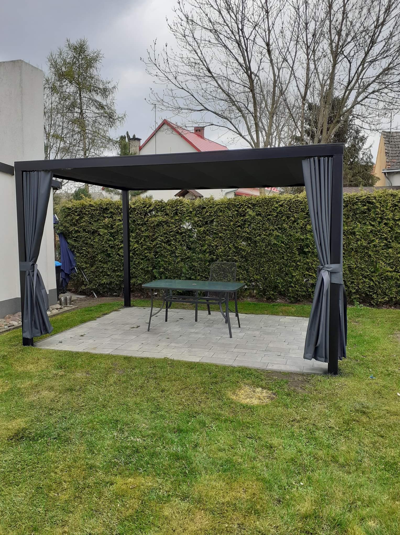Nowoczesna, czarna pergola z szarymi zasłonami na tarasie z szarej kostki brukowej, zielony trawnik i wysoki żywopłot w tle.