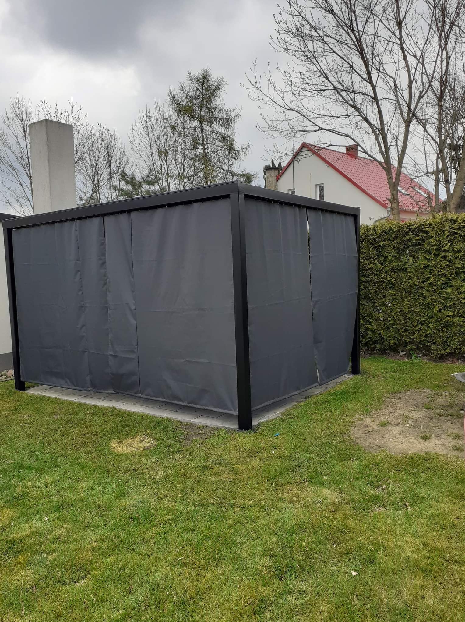 Zadaszona, czarna pergola ogrodowa z zasłonami w kolorze grafitowym, na kostce brukowej, otoczona trawnikiem i żywopłotem, z widocznym budynkiem mieszkalnym w tle.