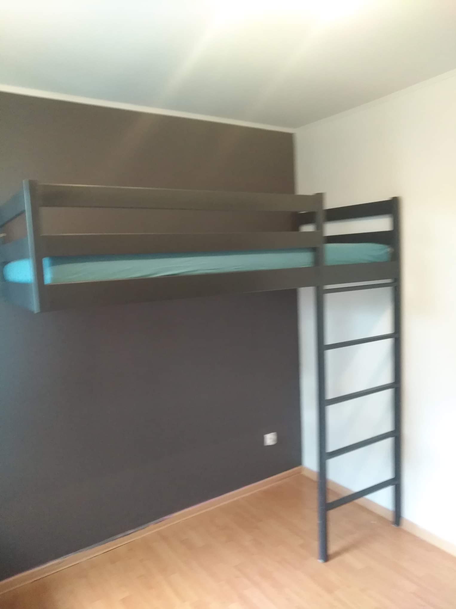 Metalowa konstrukcja łóżka na antresoli z drabinką, widok z dołu na tle ściany w dwóch odcieniach szarości i podłogi z paneli.