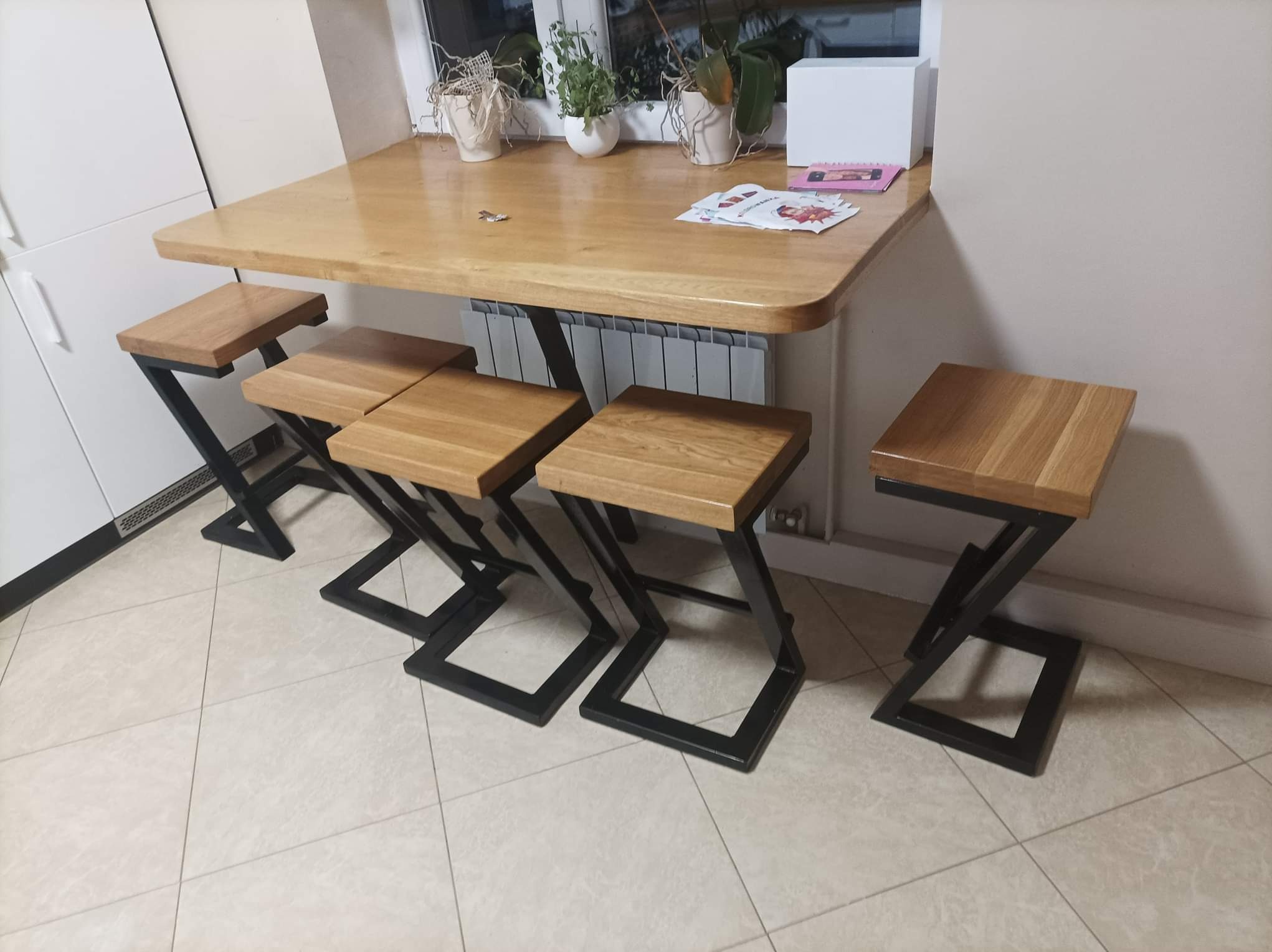 Blat dębowy przy oknie z czterema hokerami o konstrukcji metalowej w kształcie litery Z, ustawione na jasnej podłodze z płytek.