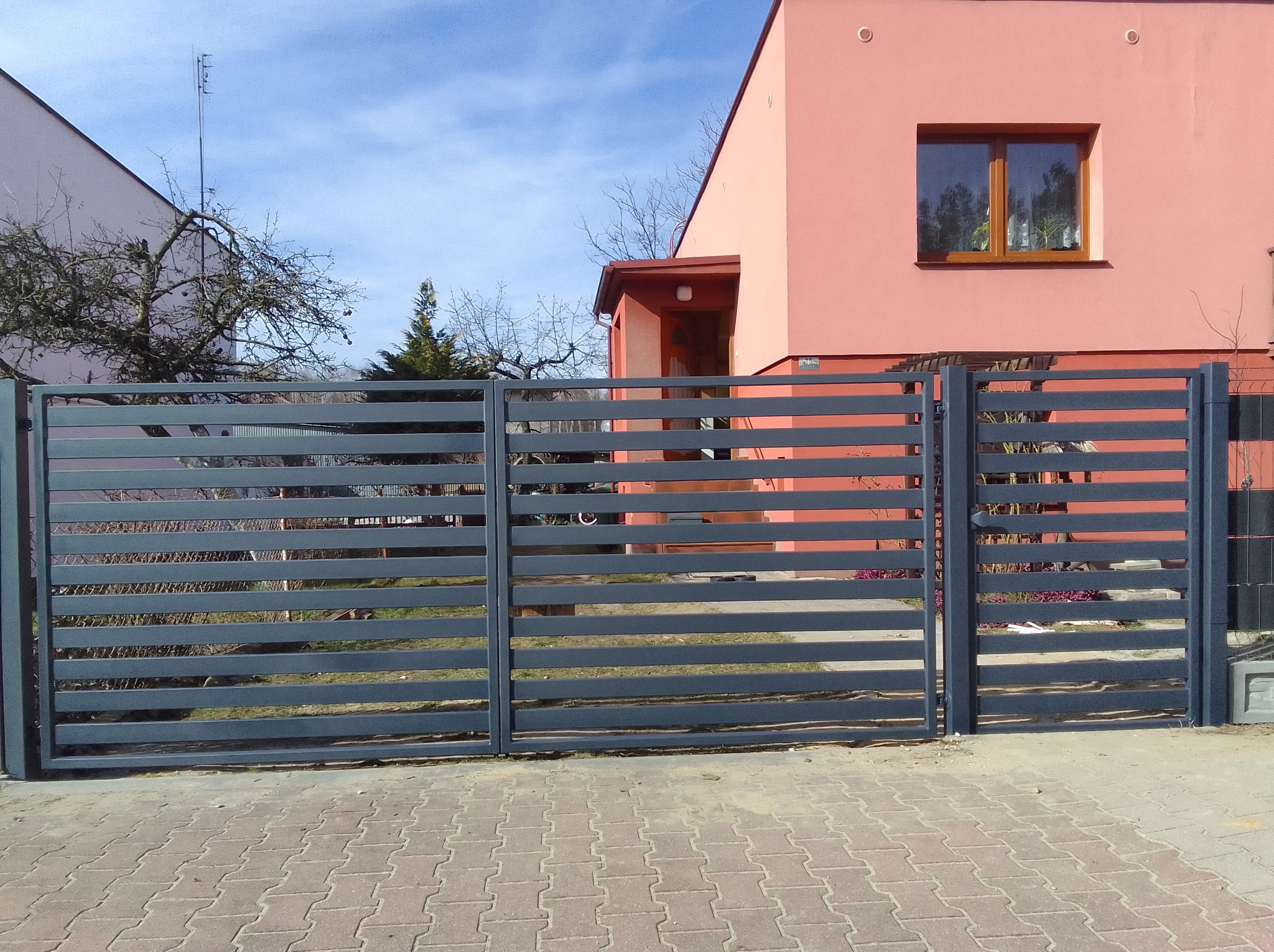 Nowoczesna, pozioma brama wjazdowa w kolorze grafitowym z furtką, na tle różowego domu jednorodzinnego, ułożona kostka brukowa.